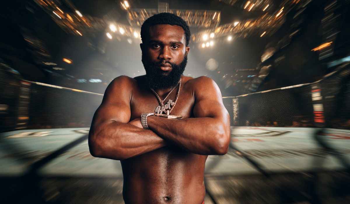 A muscular male fighter with a beard stands shirtless inside an MMA cage with arms crossed, wearing a large “Boots” chain and wristwatch under bright arena lights