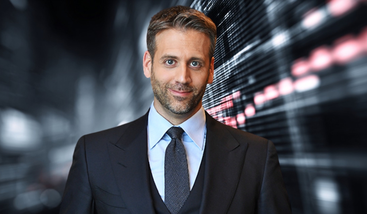 A man in a suit smiling in front of a blurred digital background with red and white lights