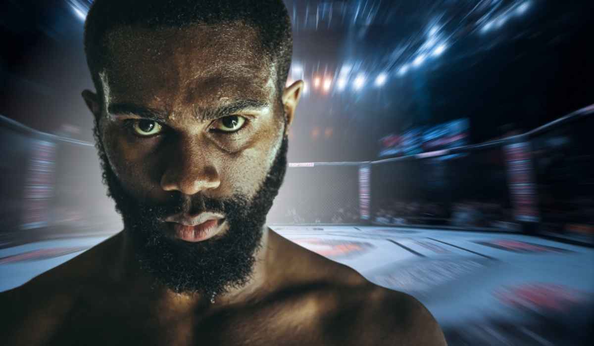 Close-up of a focused male fighter with intense eyes and a beard, standing inside a lit MMA cage before a match