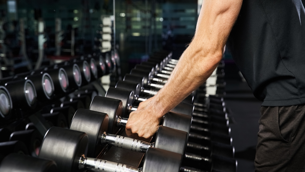 Man picks dumbbells from rack in gym, showing simple workout gear and equipment used for strength training