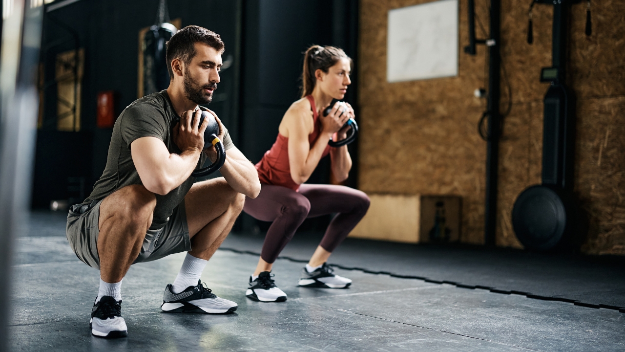 Man and woman perform squats with kettlebells in a gym, showing proper form and stable footwear